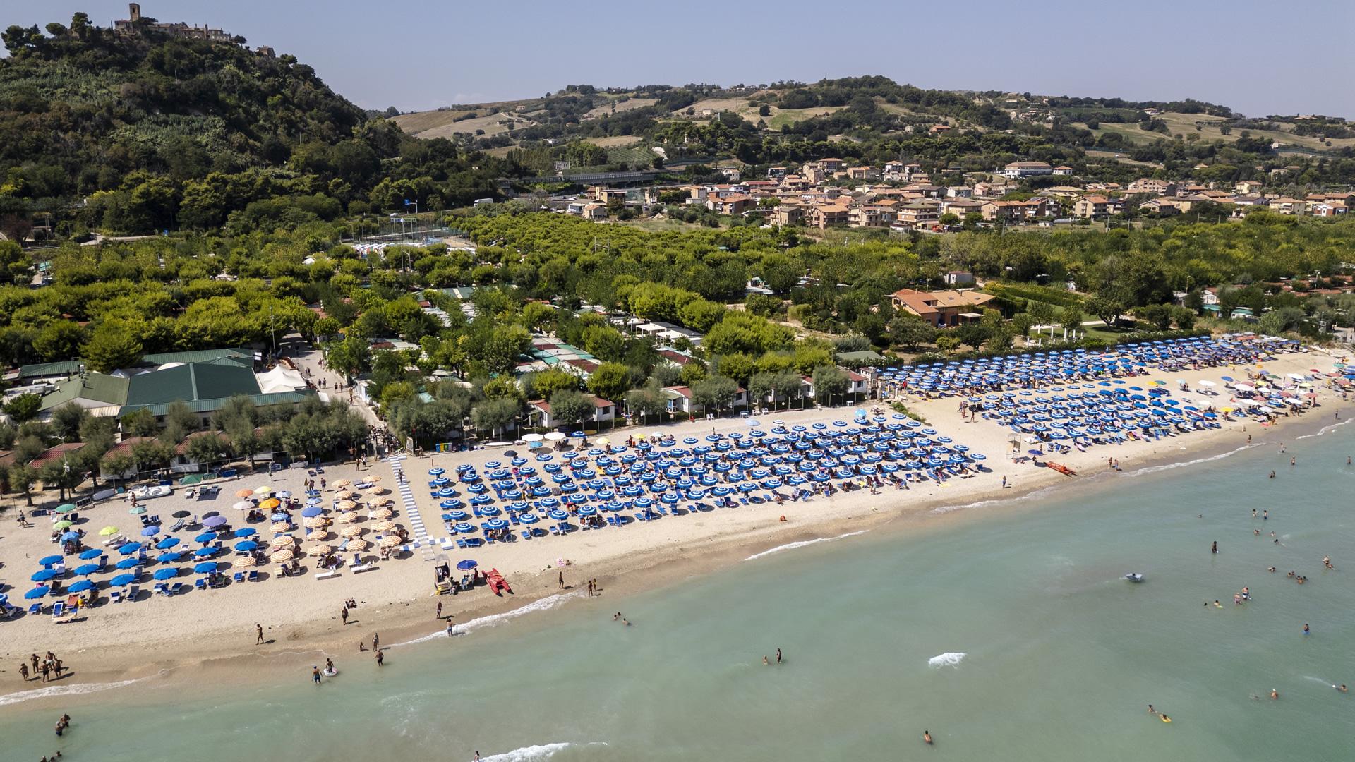 Spiaggia affollata con ombrelloni blu, vicino a colline verdi e villaggio.